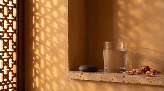 Late afternoon sun through a mashrabiya screen onto warm plaster, with a condensation-beaded glass of water and dried rose petals on a travertine ledge